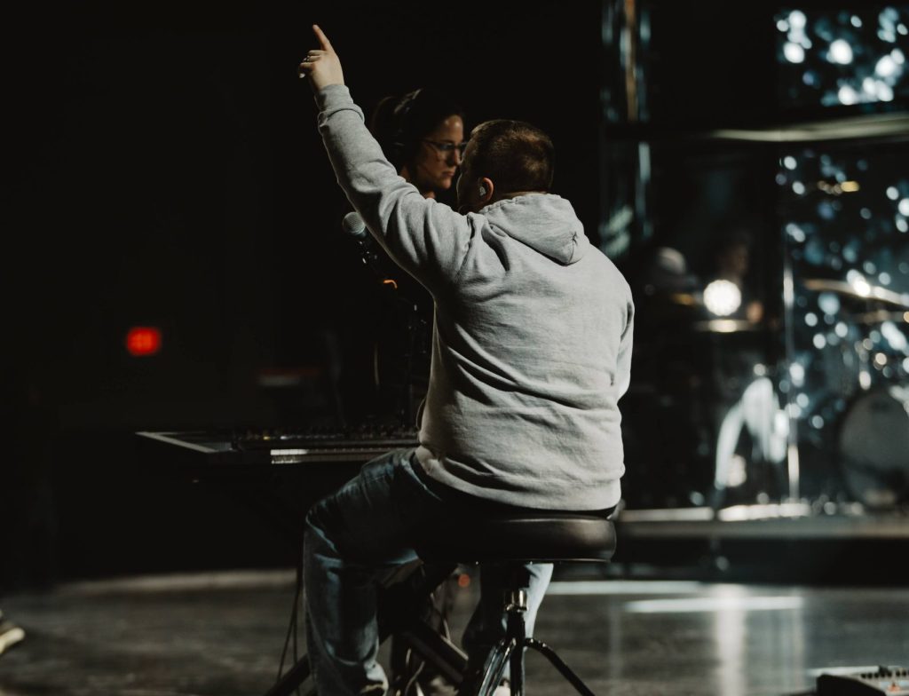 Performer in a gray hoodie sits on a stationary bike on a dimly lit stage, raising one arm as if signaling or conducting.