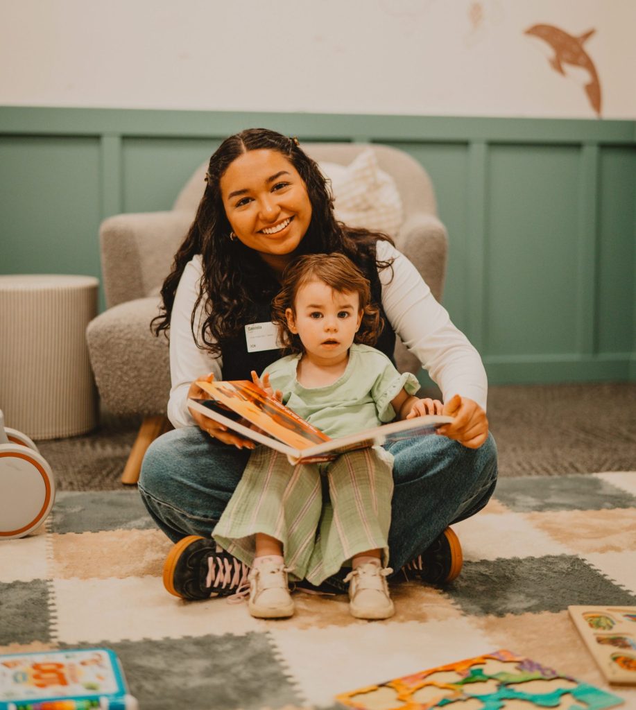 Woman and young child sitting on a rug, reading a book together in a cozy playroom with toys nearby