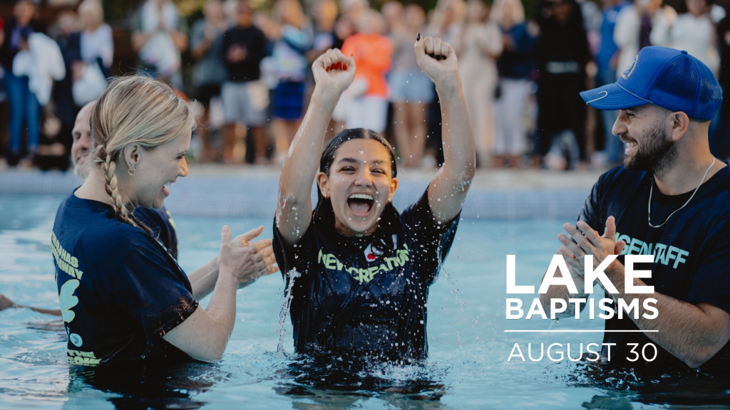 Woman in water raises arms in celebration during a baptism, crowd cheering in the background at a poolside event