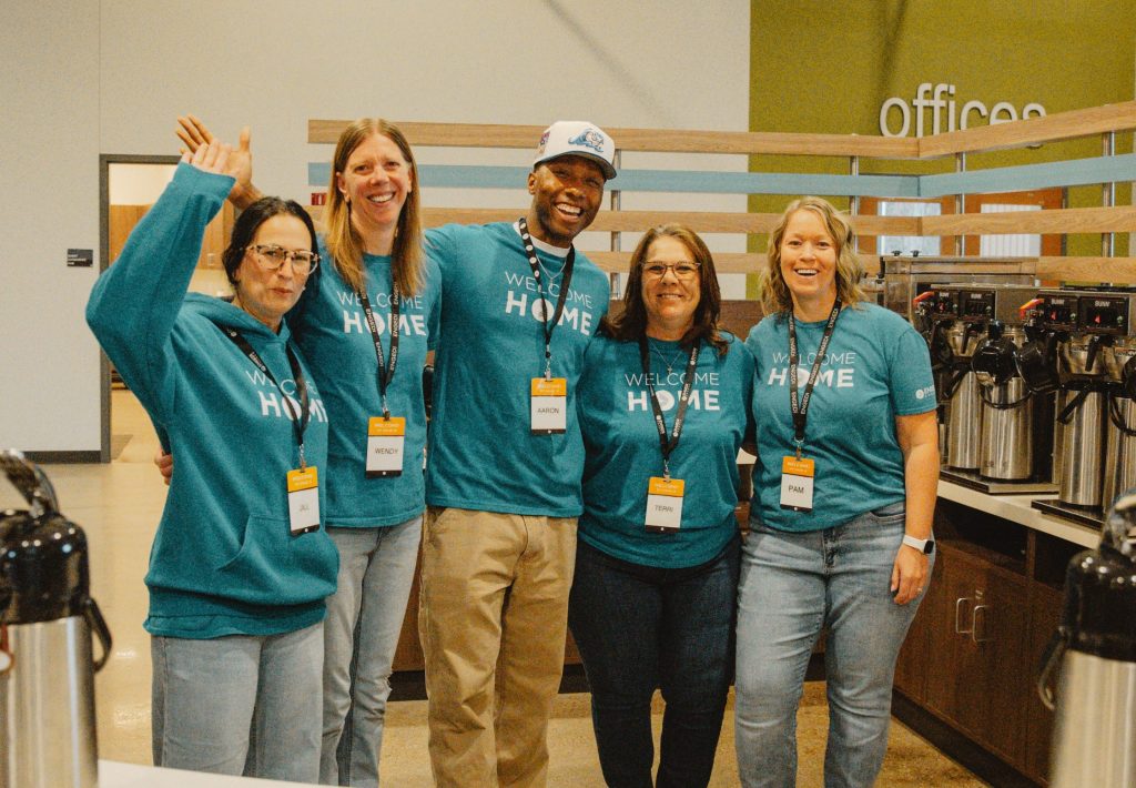 Five adults in teal 'Welcome Home' shirts pose together in a break room with coffee stations in the background, smiling and waving.
